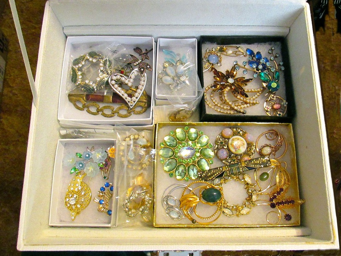 En organiserad samling vintagesmycken i fackförsedda vita lådor, som visar olika bijouterier inklusive utsmyckade broscher, pärlhalsband och nålar.